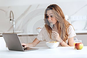 Thoughtful young woman eating and using laptop