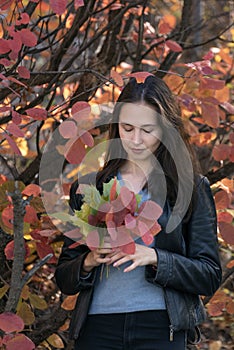 Thoughtful young woman in forest. Portrait of brunette girl with autumn leaves in her hands. Vertical frame