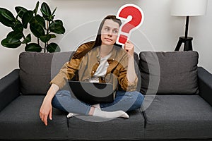 Thoughtful young woman holding red question mark, looking aside, sitting alone on couch at home. Portrait of caucasian female