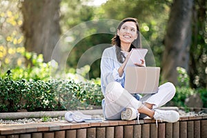 Thoughtful young woman with a laptop sitting cross-legged on a bench in a park