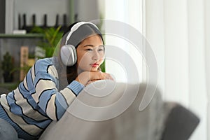 Thoughtful young woman listening to calm music in headphone and looking away while relaxing on couch
