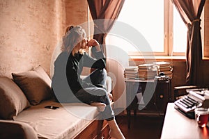 Thoughtful young woman looking through window while sitting on bed