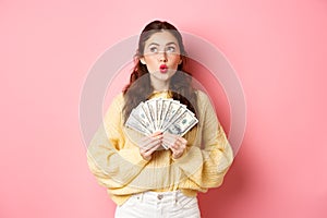 Thoughtful young woman pucker lips, looking aside with pensive face, holding dollar bills, dreaming of buying something