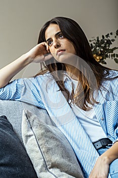 Thoughtful young woman with sad expression sitting on sofa at home looking outside and thinking bad problems on life. Lonely