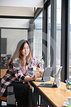 Thoughtful young woman sitting in coffee shop with her computer tablet and looking out of window.