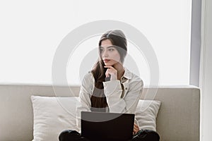 Thoughtful young woman sitting with laptop on couch