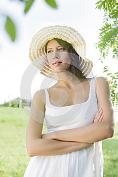 Thoughtful young woman in sundress and hat standing arms crossed in park