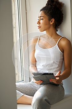 Thoughtful young woman with tablet pc looking out the window, feeling cozy while sitting alone by the window at home