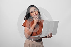 Thoughtful young woman wearing brown shirt and eyeglasses looking aside while holding laptop isolated over white background