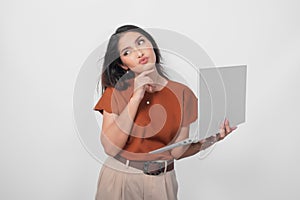 Thoughtful young woman wearing brown shirt looking aside while holding laptop  over white background