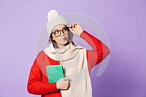 Thoughtful young woman wearing winter hat isolated over purple background reading book