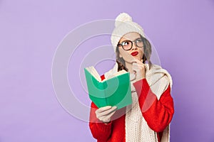 Thoughtful young woman wearing winter hat isolated over purple background reading book