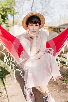 Thoughtfully green-eyed girl in trendy shoes chilling in red hammock, tired after long journey with friends. Portrait of