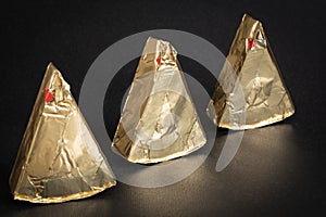 Thre triangular pieces of melted cream cheese wrapped in a golden aluminium foil on a black background.