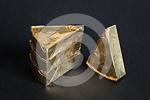 Thre triangular pieces of melted cream cheese wrapped in golden aluminium foil on black background. Portioned triangular cheeses
