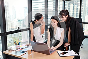 Three asian colleagues businesswomen whispering or gossiping someone from internet with laughing while working at the office with