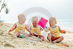 Three baby Toddler sitting on a tropical beach in Thailand and playing with sand toys. The yellow shirts. Two boys and one girl