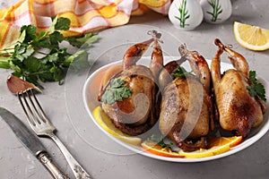 Three baked quails served with lemon, orange and parsley on a white plate on a gray background. Closeup