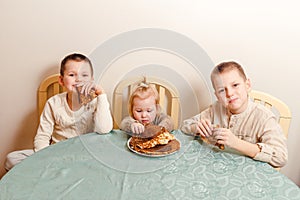Three beautiful children sit at table in the kitchen and eat large round pancakes. Carnival Festival