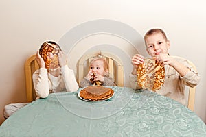 Three beautiful children sit at table in the kitchen and eat large round pancakes. Carnival Festival