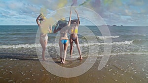 Three beautiful girls in bikinis dance happily on the beach with colored smoke fireworks using their hands to swing the colored sm