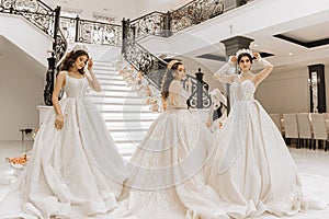 Three beautiful girls in luxurious wedding dresses with long trains pose in the hall near the steps of a restaurant