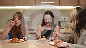 Three beautiful girls are sitting at the kitchen table and eating prepared food on the plates with a fork. The owner of