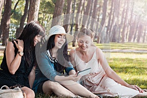 Three beautiful young women rest and joying at summer park with smartphone. Group of happy international people