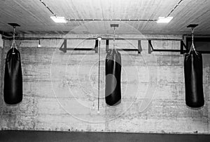 Three black punching bags in the empty boxing gym with naked grunge wall in background black and white