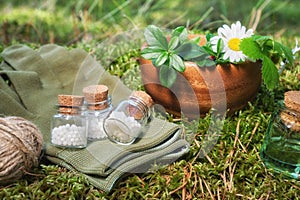 Three bottle of homeopathic globules, infusion bottle, wooden mortar of medicinal herbs, gloves on a moss in forest