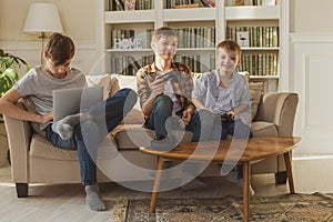 Three boys, brothers, sit on the couch at home in the sunlight and play computer and video games.