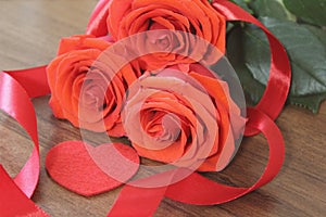 Three bright red roses, sign heart and satin ribbon on wooden background closeup.