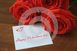 Three bright red roses and a small sheet of paper with the inscription!`Happy Valentines day!` on a wooden brown background closeu