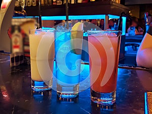 Three brightly colored cocktails stand on the bar counter. Each drink is in its own glass, from bright red to soft blue. The photo