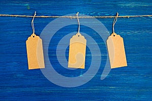 Three brown blank paper price tags or labels set hanging on a rope on the blue background.