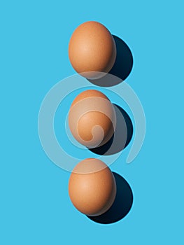 Three brown chicken eggs lie on top of each other on a blue background. Bright light, deep shadow, vertical