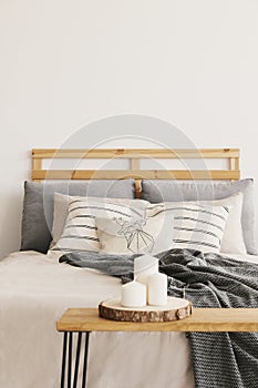 Three candles on the wooden table next to cozy bed with patterned pillows and grey blanket, real photo