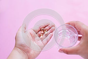 Three capsules of omega three fish oil in one hand and a glass of water in the other on a pink background