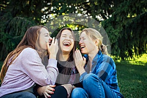 Three cheerful girls whisper and fun against green foliage in the park. Women joke and laugh, close up. Students rest outdoor