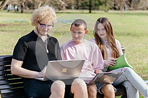 Three cheerful students study and work together outdoors on a sunny day, using laptops and notebooks while sitting on a