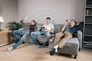 Three cheerful young guys use phones on a soft couch.