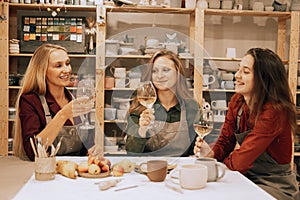 Three cheerful young women friends are having fun in a ceramic workshop. They drink wine and joke, have fun.