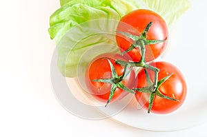 Three cherry tomatoes with stems, green lettuce leaves and a white plate, isolated on white tabletop background surface, top view