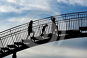 Three children as silhouettes climbing up the steps of a large metal bridge against a blue sky with clouds, future challenge for