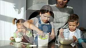 Three children of different ages knead the dough. Elder sister to the flour in a bowl. Mom in the background manages the