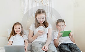 Three children of different ages are sitting on a bed with different electronic gadgets. A little girl sits with a laptop, a