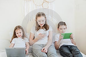 Three children of different ages are sitting on a bed with different electronic gadgets. A little girl sits with a laptop, a