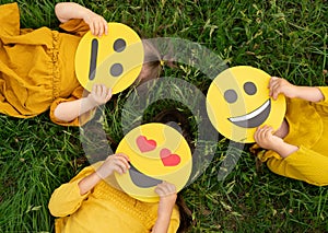 Three children lying on the grass are holding cardboard emoticons