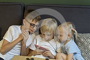 Three children sit on couch and look at the phone. Siblings enthusiastically look at screen of smartphone. Modern children