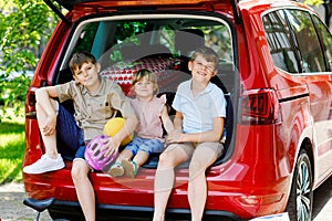 Three children, two boys and preschool girl sitting in car trunk before leaving for summer vacation with parents. Happy